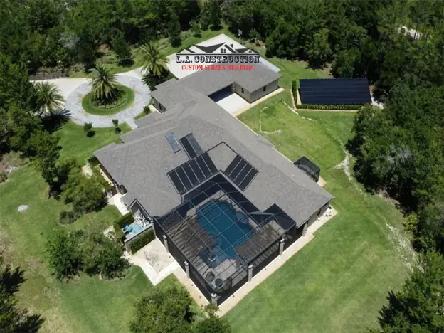 Aerial view of a home with a mansard-style pool screen enclosure, solar panels and a wooded lot